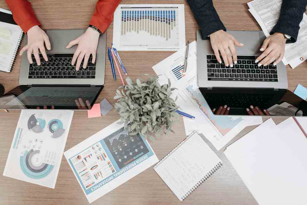 Overhead view of two people working on a franchise business plan with laptops, financial charts, graphs, and papers spread across a desk.